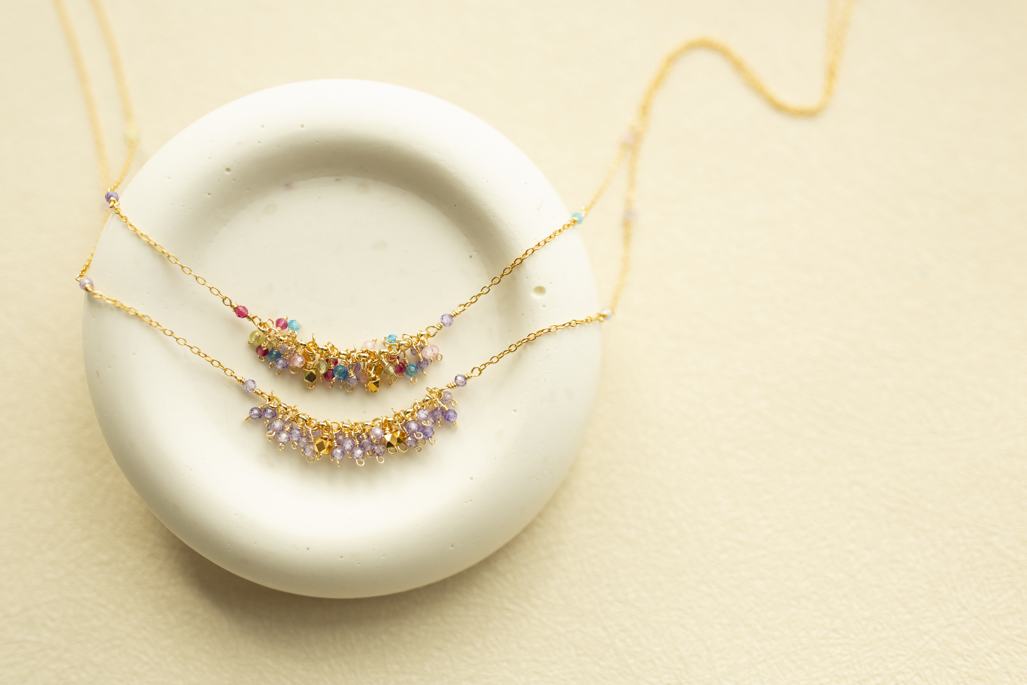 Two delicate gold chain necklaces with small multicolored beads and crystal-like accents, displayed on a round white dish against a warm beige background.