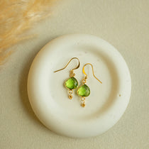 A pair of gold-tone dangle earrings with green faceted gemstone drops, displayed on a round white ceramic dish against a warm beige textured background.