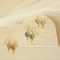 Three pairs of earring in gold with blue, green and pink gemstones on a beige background