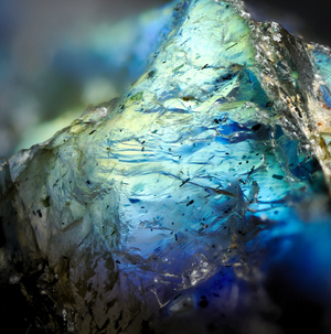 A close-up macro shot of a translucent blue and green mineral or crystal specimen with iridescent tones and small dark inclusions.